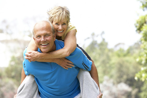 Older man and his female partner smile as she rides piggy-back with her arms around his shoulders.